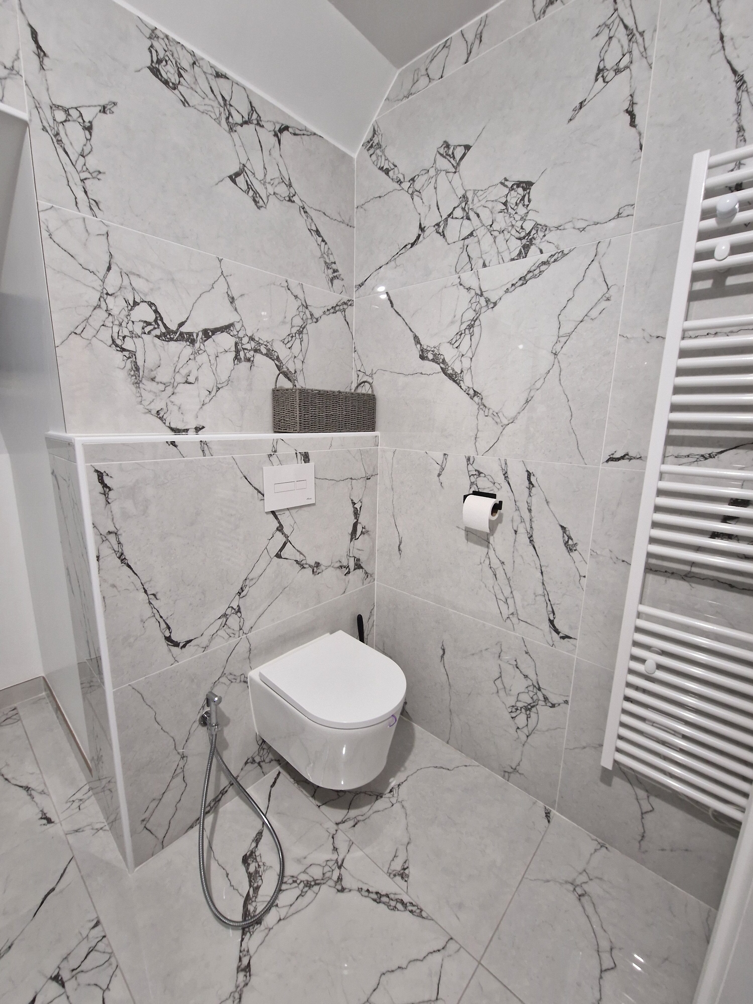 Salle de bain moderne avec WC suspendu, carrelage effet marbre blanc et noir, douchette de bidet et sèche-serviettes blanc.