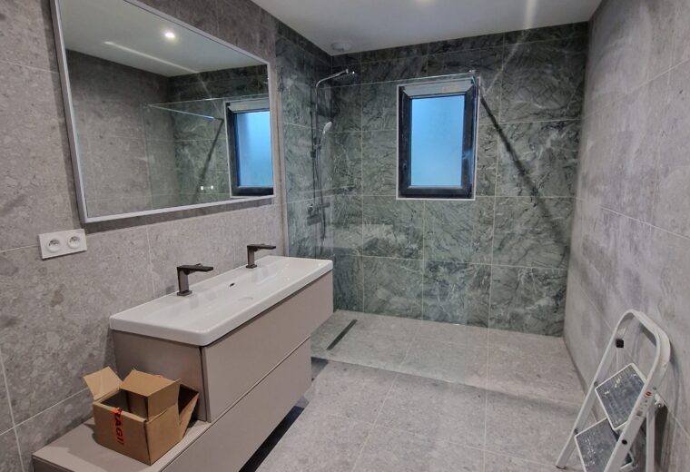 Salle de bain moderne en cours de rénovation avec carrelage gris clair et mural effet marbre vert, meuble double vasque blanc et beige, grand miroir rectangulaire, douche à l'italienne et deux fenêtres.