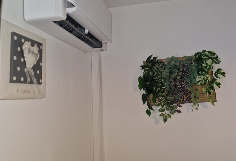 Mur blanc avec climatiseur mural Daikin, cadre décoratif en forme de cœur, rideau noir avec embrasse en bois, et jardinière murale avec des plantes vertes.