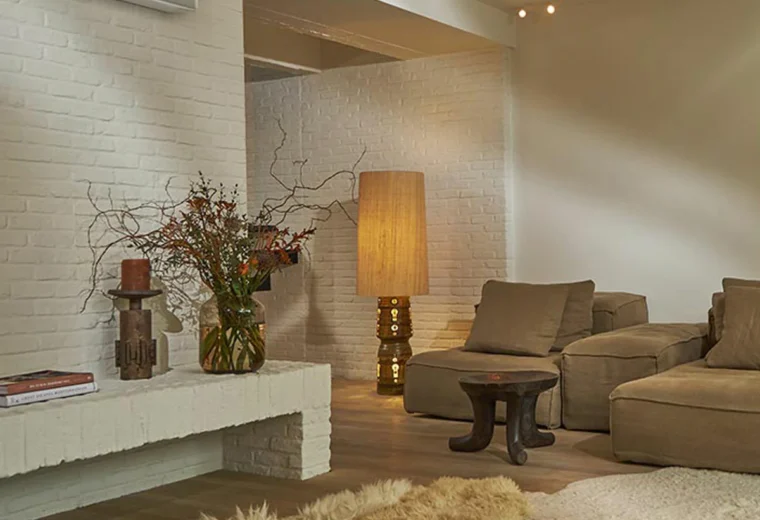 Salon au style bohème avec mur en briques peintes, climatiseur mural blanc, banquette décorative, lampe de sol originale et canapé modulaire couleur terre, un tapis en fausse fourrure au sol.