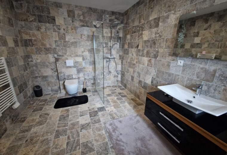 Salle de bain moderne entièrement carrelée en pierre naturelle aux tons bruns et gris, avec douche à l'italienne, WC suspendu, sèche-serviettes blanc, et meuble-lavabo noir avec vasque blanche et grand miroir.