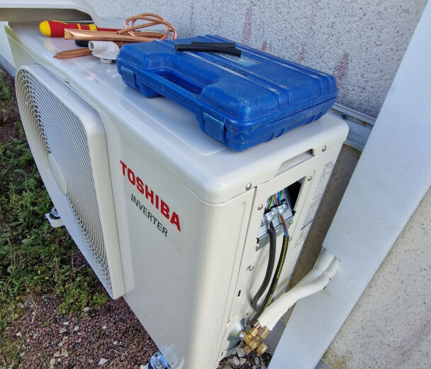 Unité extérieure de climatisation Toshiba Inverter en cours d'installation ou maintenance, avec outils et câblage visible, posée au sol contre un mur extérieur.