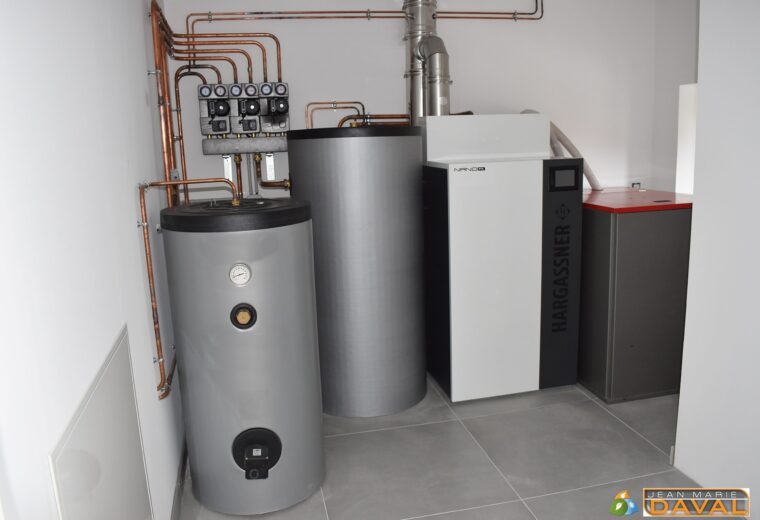 Un local technique moderne et propre, doté d'un système de chauffage complet Hargassner Nano-PK à granulés de bois. L'installation comprend la chaudière blanche et noire, deux grands ballons tampons/d'eau chaude argentés, et des tuyauteries en cuivre méticuleusement agencées sur un mur blanc.