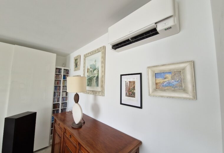 Intérieur moderne et lumineux avec climatiseur mural blanc Daikin au-dessus d'une enfilade en bois foncé, ornée d'une lampe de table design et de tableaux encadrés sur un mur blanc. Une bibliothèque et un meuble blanc sont visibles en arrière-plan.