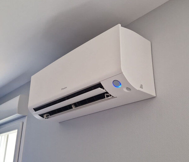 Climatiseur mural blanc Daikin avec lumière bleue, installé sur un mur gris clair près d'une fenêtre.