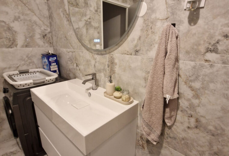 Salle de bain moderne carrelée effet marbre beige-gris, avec meuble sous-vasque blanc, vasque et miroir rond lumineux, machine à laver, panier à linge et poubelle.