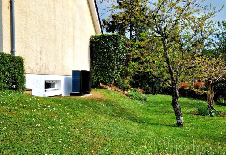 Maison individuelle à façade beige clair et toit en tuiles, bordée par un grand jardin verdoyant parsemé de marguerites. Un arbre au jeune feuillage printanier et une unité extérieure (pompe à chaleur) sont visibles sur le terrain.