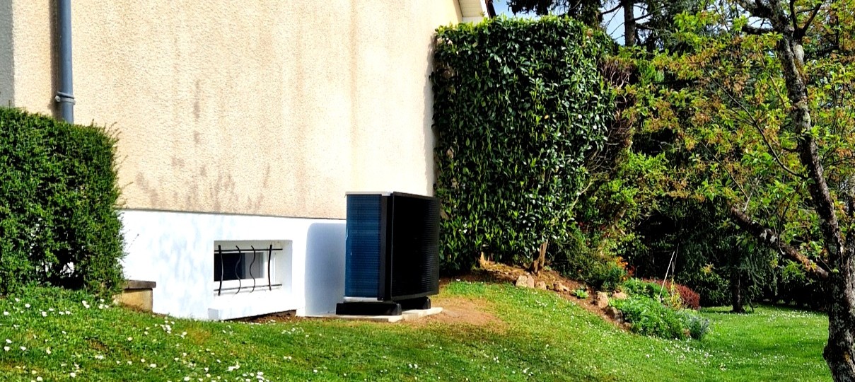 Maison individuelle à façade beige clair et toit en tuiles, bordée par un grand jardin verdoyant parsemé de marguerites. Un arbre au jeune feuillage printanier et une unité extérieure (pompe à chaleur) sont visibles sur le terrain.
